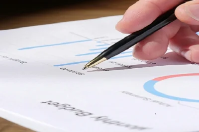 A hand holding a pen pointing at a monthly budget chart with bar graphs and pie chart